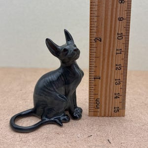 K&ouml;nnte beinhalten: Eine schwarze Keramikfigur eines haarlosen Katers, der mit seinem Schwanz um seine Beine gewickelt sitzt. Die Figur ist etwa 5 cm hoch und befindet sich neben einem Lineal zur Gr&ouml;&szlig;enbestimmung.