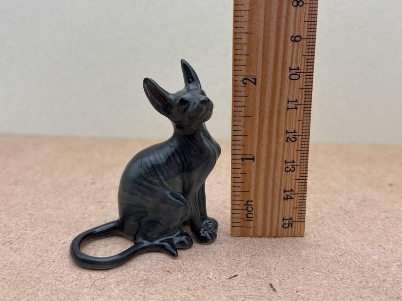 K&ouml;nnte beinhalten: Eine schwarze Keramikfigur eines haarlosen Katers, der mit seinem Schwanz um seine Beine gewickelt sitzt. Die Figur ist etwa 5 cm hoch und befindet sich neben einem Lineal zur Gr&ouml;&szlig;enbestimmung.