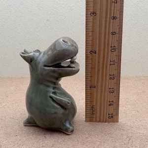 K&ouml;nnte beinhalten: Eine Keramikfigur eines grauen Flusspferdes mit offenem Maul, stehend auf einer hellbraunen Oberfl&auml;che. Das Flusspferd ist etwa 10 cm hoch.