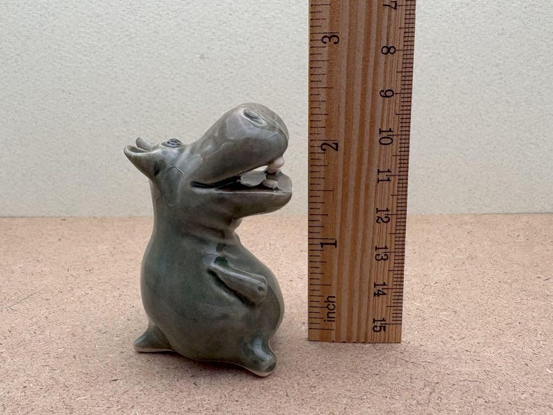 K&ouml;nnte beinhalten: Eine Keramikfigur eines grauen Flusspferdes mit offenem Maul, stehend auf einer hellbraunen Oberfl&auml;che. Das Flusspferd ist etwa 10 cm hoch.