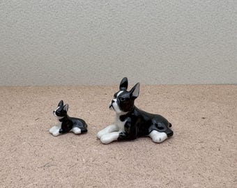 May include: Two black and white ceramic Boston Terrier figurines. The larger figurine is lying down with its head turned to the side. The smaller figurine is sitting up with its head tilted to the side.