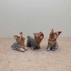 Puede incluir: Tres figuritas de cer&aacute;mica de perros Yorkshire Terrier. Los perros est&aacute;n en varias poses, uno mirando hacia arriba, otro mirando hacia adelante y otro mirando hacia un lado. Las figuritas est&aacute;n pintadas en tonos de marr&oacute;n, negro y gris.