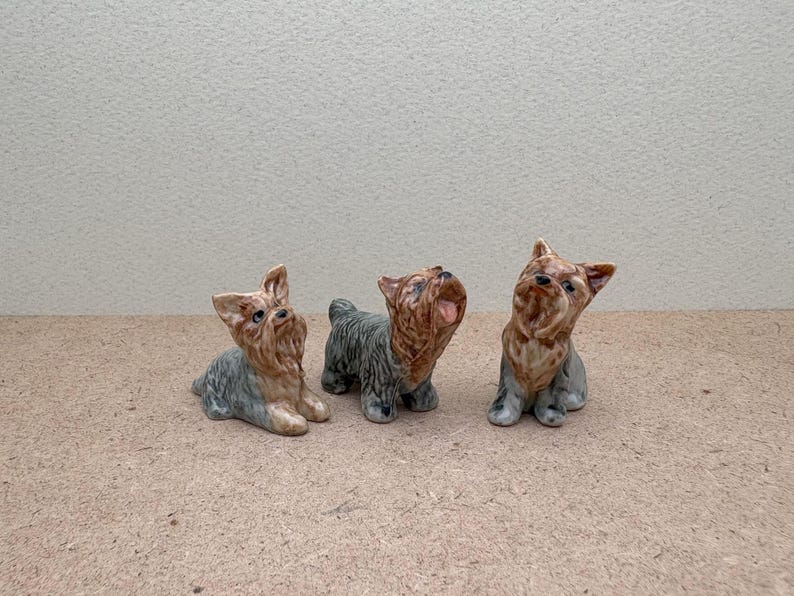 Puede incluir: Tres figuritas de cer&aacute;mica de perros Yorkshire Terrier. Los perros est&aacute;n en varias poses, uno mirando hacia arriba, otro mirando hacia adelante y otro mirando hacia un lado. Las figuritas est&aacute;n pintadas en tonos de marr&oacute;n, negro y gris.