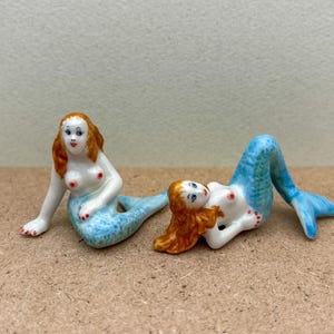 Peut inclure: Deux figurines de sirène en porcelaine avec des queues bleues et des cheveux roux. Une sirène est assise avec sa queue enroulée derrière elle, tandis que l'autre est allongée sur le côté avec sa queue étendue.