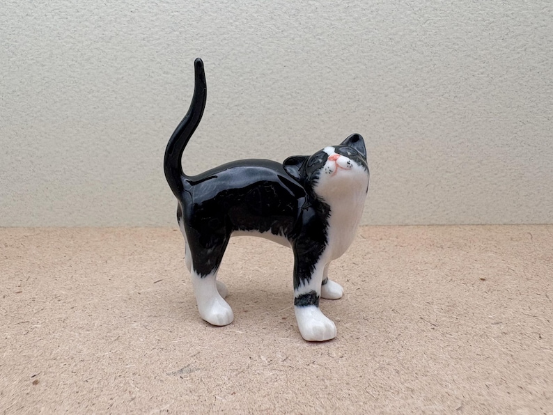 K&ouml;nnte beinhalten: Keramikfigur einer Katze im Tuxedo-Muster, mit gl&auml;nzendem Schwarz-Wei&szlig;-Finish. Die Katze steht mit nach oben geneigtem Kopf und gew&ouml;lbtem Schwanz. Die Pfoten der Katze sind wei&szlig;, die Nase rosa. Dekoration.