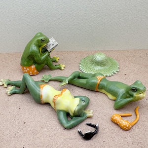 May include: Three ceramic frog figurines on a brown surface. One frog is sitting and reading a book, another is lying on its back with its arms behind its head, and the third is lying on its stomach with its head resting on its arms. All three frogs are wearing bathing suits and have green and orange polka dots.
