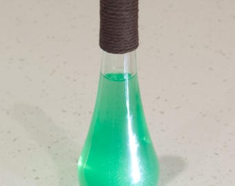 Puede incluir: Una botella de vidrio transparente con un líquido verde en su interior. La botella tiene una cuerda marrón envuelta alrededor del cuello y una tapa negra.