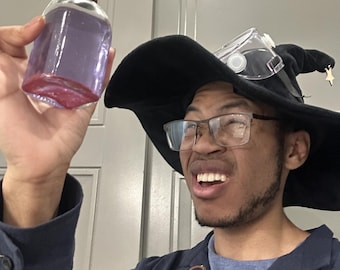 Puede incluir: Una persona que lleva un sombrero de bruja negro con una pantalla facial transparente y gafas sostiene una botella de vidrio transparente con un líquido morado y un tapón de corcho marrón.
