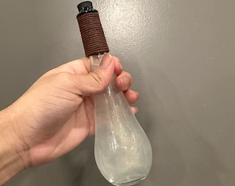 Puede incluir: Una botella de vidrio transparente con una cuerda marrón envuelta alrededor del cuello. La botella está llena de un líquido transparente y tiene un efecto de brillo plateado.