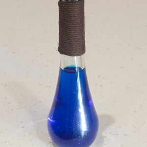 Könnte beinhalten: Eine klare Glasflasche in Tropfenform, gefüllt mit einer leuchtend blauen Flüssigkeit. Die Flasche hat eine braune Schnur um den Hals gewickelt.