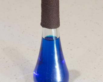 Puede incluir: Una botella de vidrio transparente con forma de lágrima llena de un líquido azul brillante. La botella tiene una cuerda marrón envuelta alrededor del cuello.