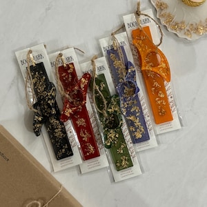 Puede incluir: Cinco marcapáginas coloridos con detalles de pan de oro. Cada marcapáginas está hecho de resina y tiene un diseño único. Los marcapáginas están empaquetados en fundas de plástico transparente con el texto "BOOKMARK" y "HANDMADE WITH LOVE BY LEAPTHINKING" impreso en ellos.