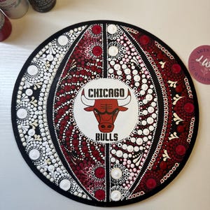 May include: A round, black, wooden plaque with a red, white, and black Chicago Bulls logo in the center. The plaque is decorated with red, white, and black dots in a mandala pattern.