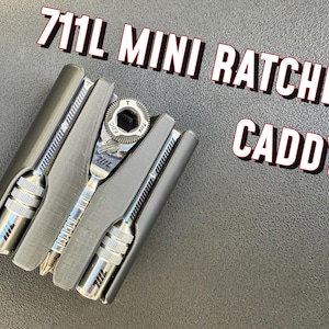May include: A 711L Mini Ratchet Caddy featuring a dark gray holder containing a silver metal ratchet tool and two silver metal screwdriver bits. The text 711L MINI RATCHET CADDY is printed in large white letters with a black outline above the tool set.