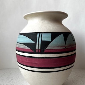 May include: White ceramic vase with a geometric design in black, blue, and pink. The vase has a rounded body and a wide opening.