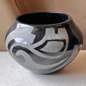 May include: A black ceramic pot with a glossy finish. The pot features a gray and black geometric design around the exterior. The pot has a wide opening and a rounded body. The design includes curved and angular shapes.