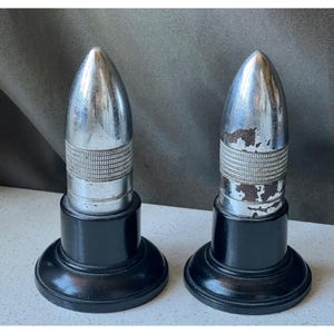 May include: Two vintage bullet-shaped bottle stoppers. The stoppers have a silver-colored, bullet-like top with a textured band and sit on a black, round base. One stopper shows signs of wear.