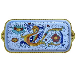 May include: Rectangular ceramic serving tray with a light blue background and a yellow border. The tray features a colorful design of a bird, flowers, and ornate scrollwork in yellow, blue, red, and green. The tray has two handles.