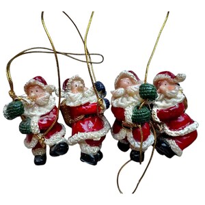 May include: Four red and white Santa Claus ornaments hanging from gold cords. Each Santa is holding a green and white gift bag and is wearing a white fur trim hat.