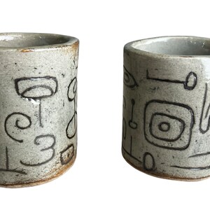 May include: Two small, white ceramic mugs with black abstract designs. The mugs have unique, angular handles.