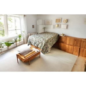 May include: A bedroom with a white bed with a green and floral patterned duvet cover. The bed is next to a wooden dresser with multiple drawers. There is a wooden stool in the foreground.