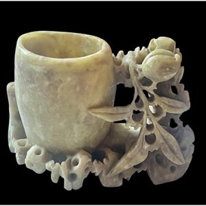 May include: A carved, light-yellow soapstone cup with intricate floral and leaf detailing. The cup has a cylindrical shape with a rounded base and is set against a black background. The carving includes a handle and decorative elements.