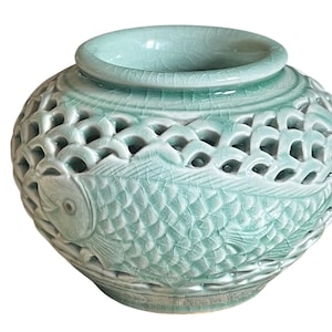 May include: A light green ceramic vase with a celadon glaze and a crackle finish. The body features an intricate openwork lattice pattern and a relief design of a fish with detailed scales wrapping around the exterior of the rounded vessel.