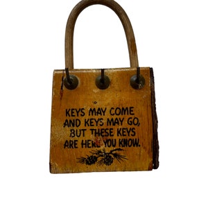 May include: A wooden key holder shaped like a small purse with a brown wooden handle. The holder has three hooks and a black printed message that reads "KEYS MAY COME AND KEYS MAY GO, BUT THESE KEYS ARE HERE YOU KNOW."