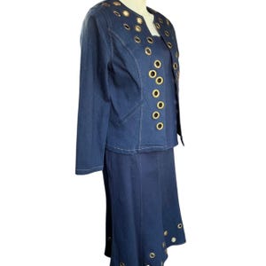 May include: A denim skirt suit in a medium wash. The jacket and skirt feature gold-tone grommet details. The jacket has a tailored fit with long sleeves and a notched collar. The skirt is a midi length.