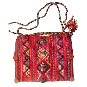 May include: A red woven shoulder bag with a geometric pattern in various colors. The bag has a braided strap and a tassel. Small pom-poms are attached to the bottom of the bag. The bag is made of a textured material.