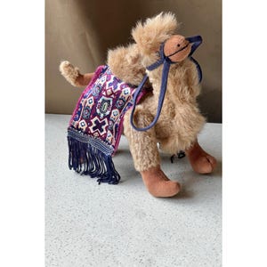 May include: A plush camel toy with a brown and white patterned saddle and a blue bridle. The camel is sitting on a white surface.