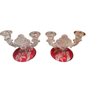 May include: A pair of two-armed candle holders made of clear glass with a central fan-shaped detail. The circular bases are colored in a deep red with a white etched floral pattern. Each holder features two candle cups on curved glass arms.