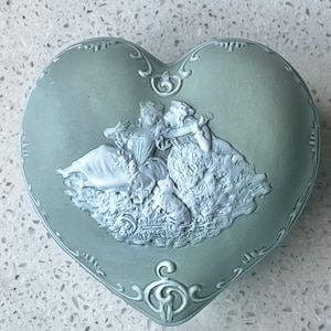 May include: A heart-shaped trinket box in a muted green hue. The lid features a white bas-relief scene of two figures in flowing gowns, a small animal, and foliage. Decorative scrollwork adorns the edges.