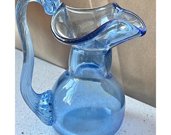 May include: A small, light blue glass pitcher with a curved handle and a flared rim. The pitcher has a rounded body and is made of transparent glass. The handle curves gracefully from the body to the top of the pitcher.