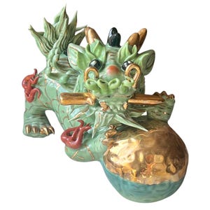 May include: A ceramic figurine of a mythical creature, primarily in shades of green, with gold and red accents. The creature has a dragon-like appearance, holding a gold sphere. The figurine is detailed with intricate designs and features.