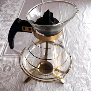 May include: A glass coffee pot with a gold rim and a black handle. The pot has a gold-trimmed base with three legs.