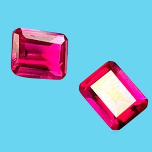 Op de afbeelding: Twee stralende, rechthoekig geslepen, felroze edelstenen tegen een lichtblauwe achtergrond. De edelstenen hebben een gepolijst, reflecterend oppervlak, dat hun interne facetten laat zien. De edelstenen hebben een levendige, verzadigde kleur.