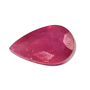 Puede incluir: Una gema de rubí facetada en forma de pera. La gema es de un color rojo rosado intenso y saturado con una superficie pulida. La piedra tiene algunas inclusiones y está sobre un fondo blanco.