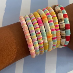 May include: A stack of colourful bracelets made of small, round beads. The bracelets are layered on a person's wrist and feature a variety of pastel colours, including pink, blue, yellow, and green.