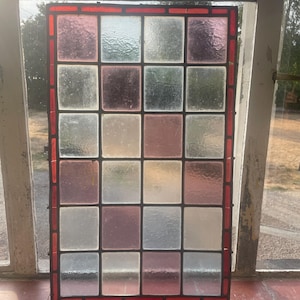 Könnte beinhalten: Ein rechteckiges Buntglasfenster-Paneel mit einem Gitter aus quadratischen Glasscheiben in verschiedenen Nuancen von klar, weiß und zartrosa. Das gesamte Paneel ist von einem Rand aus kleinen, rechteckigen roten Glasstücken in dunklem Metall gefasst.