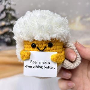 May include: A crocheted beer mug with a white foam top and a yellow body. The mug has a smiling face and is holding a card that says "Beer makes everything better."