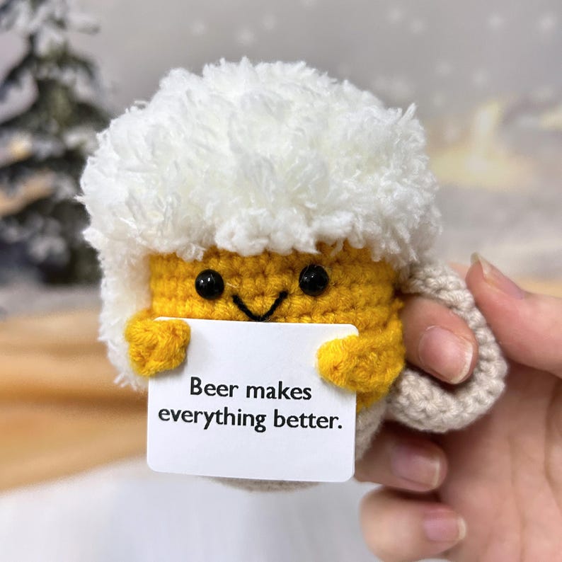 May include: A crocheted beer mug with a white foam top and a yellow body. The mug has a smiling face and is holding a card that says "Beer makes everything better."