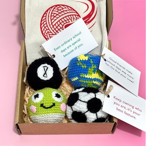 May include: A cardboard gift box containing four crocheted spheres: a black eight-ball, a blue and yellow globe, a green frog face, and a black and white soccer ball. Three white paper tags with printed text are attached to the items with twine.