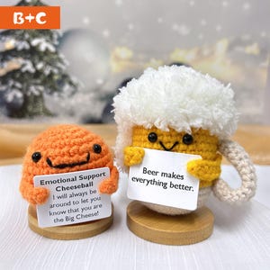 May include: Two crocheted plushies holding signs. The orange plushie is shaped like a ball and says "Emotional Support Cheeseball I will always be around to let you know that you are the Big Cheese!" The yellow plushie is shaped like a beer mug and says "Beer makes everything better."