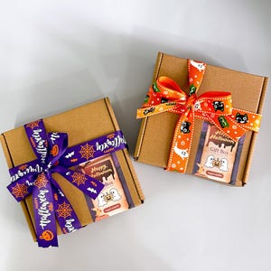 May include: Two brown cardboard gift boxes tied with Halloween-themed ribbons. One box has a purple ribbon with orange text and spiderweb designs. The other has an orange ribbon with black cat and ghost designs. Each box has a "Happy Halloween Gift Box" label.