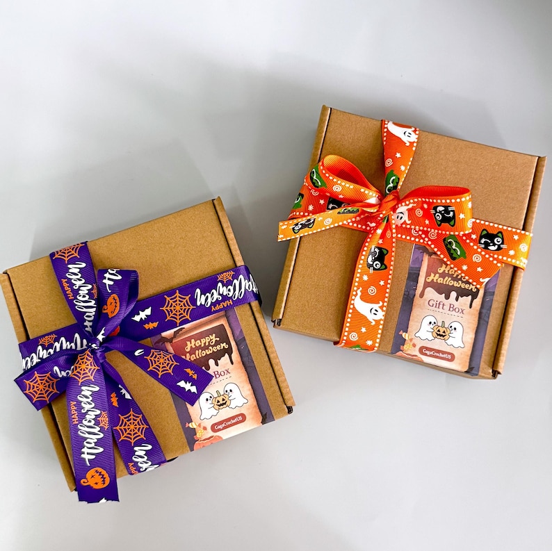 May include: Two brown cardboard gift boxes tied with Halloween-themed ribbons. One box has a purple ribbon with orange text and spiderweb designs. The other has an orange ribbon with black cat and ghost designs. Each box has a "Happy Halloween Gift Box" label.