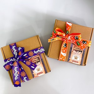 K&ouml;nnte beinhalten: Zwei braune Geschenkboxen aus Pappe, jeweils mit einem dekorativen Band gebunden. Eine Box hat ein lila Band mit dem Text "Happy Halloween" und Spinnennetz- und Fledermausgrafiken. Die andere hat ein orangefarbenes Band mit Halloween-Motiven.