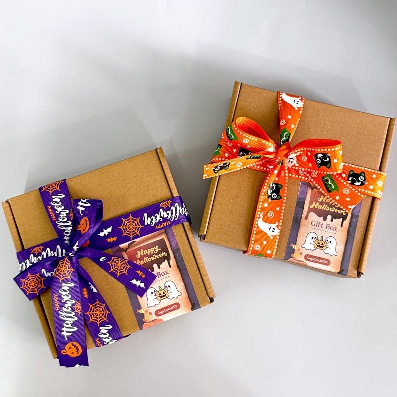 K&ouml;nnte beinhalten: Zwei braune Geschenkboxen aus Pappe, jeweils mit einem dekorativen Band gebunden. Eine Box hat ein lila Band mit dem Text "Happy Halloween" und Spinnennetz- und Fledermausgrafiken. Die andere hat ein orangefarbenes Band mit Halloween-Motiven.