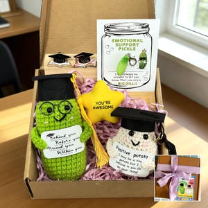 May include: A gift box containing graduation-themed items. Features crocheted pickle and potato figures wearing mortarboards, a yellow star, and a card that reads "Emotional Support Pickle". Also includes pins with the graduation year.