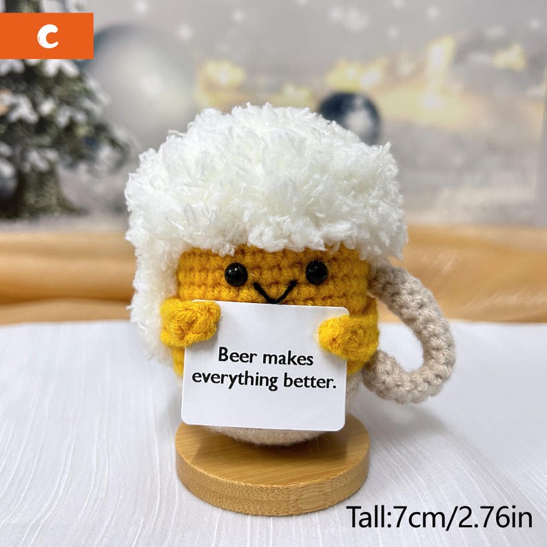 May include: A crocheted beer mug with a white fuzzy top, holding a sign that says "Beer makes everything better." The mug is standing on a wooden base. Tall: 7cm/2.76in